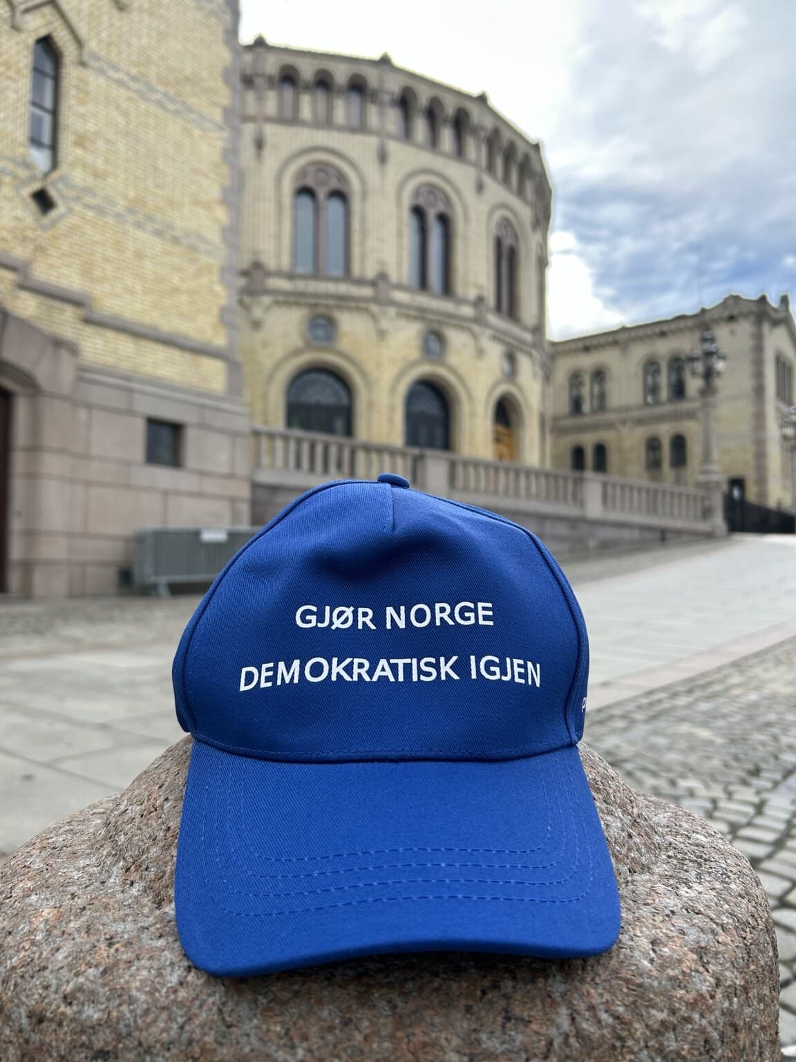 Blå caps med teksten «Gjør Norge demokratisk igjen», med Stortinget i bakgrunnen.
