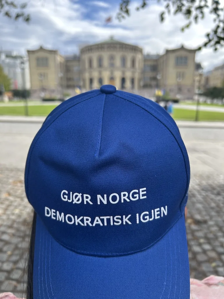 Blå caps med teksten «Gjør Norge demokratisk igjen», med Stortinget i bakgrunnen.